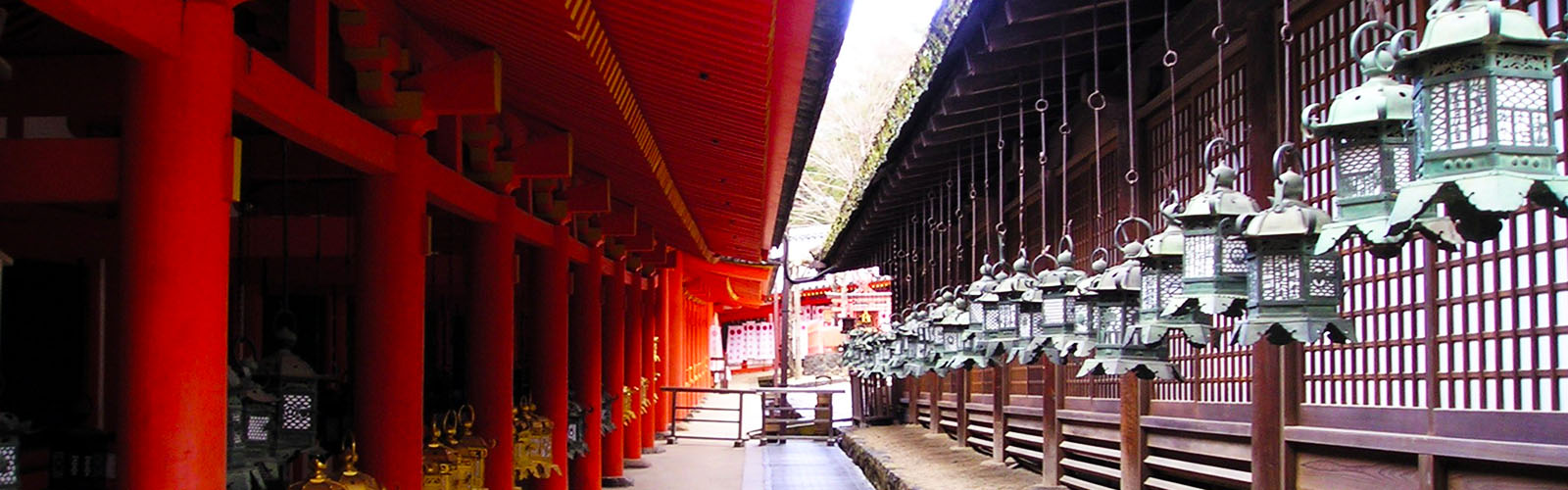 A narrow walkway with a bright red building with pillars on the left, and a muted purple building on the right with hanging green lanterns. 